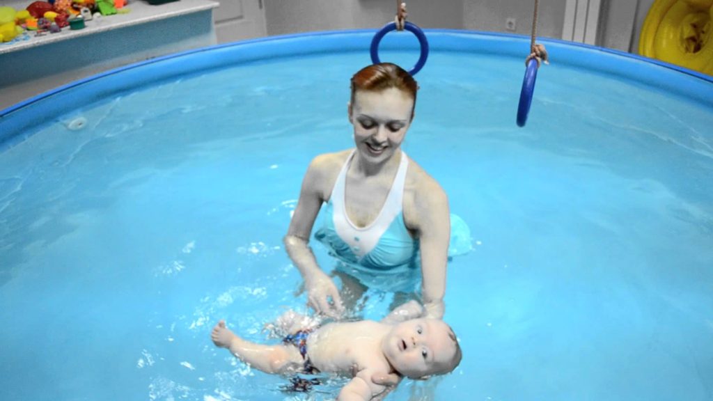 Девушка держит на руке малыша, лежащего на воде на спине, над бассейном брусья